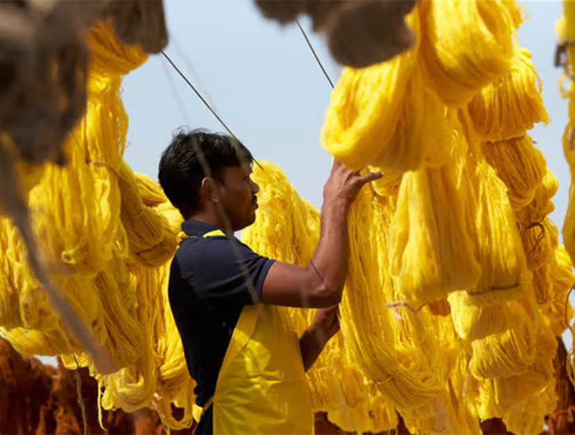 A man in a yellow apron arranging bundles of bright yellow dyed yarn hanging to dry on bamboo racks outdoors.