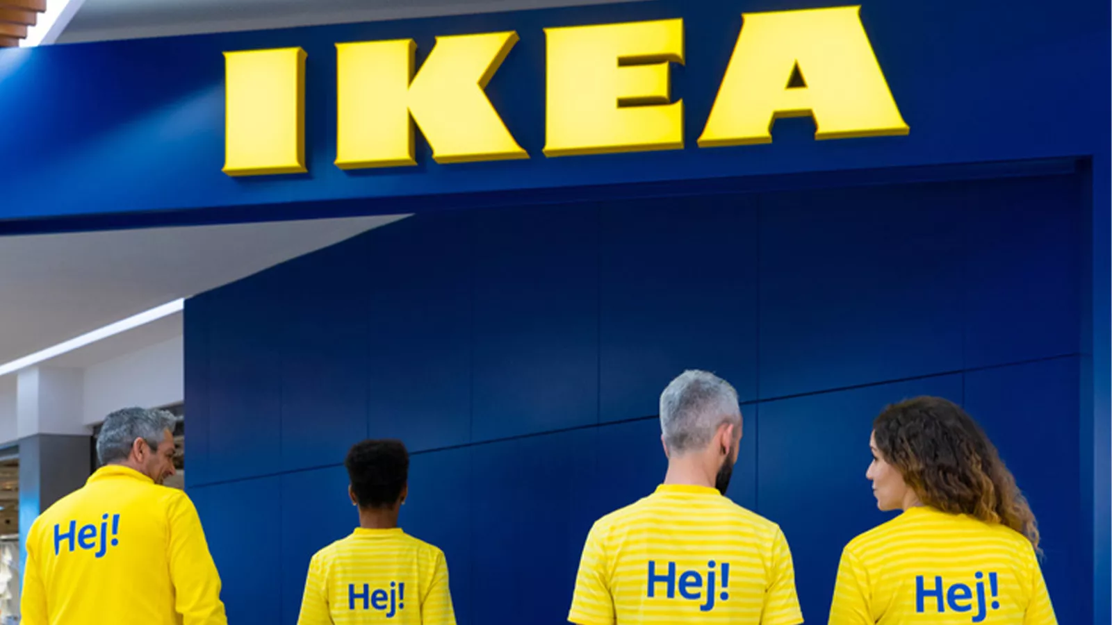 Four employees wearing yellow "Hej!" shirts standing beneath the IKEA store logo.